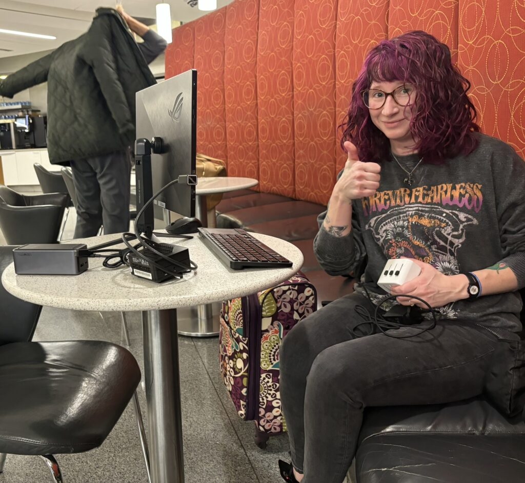 A person with long purple hair gives a thumbs up at a table with a full keyboard, monitor and tiny computer. 