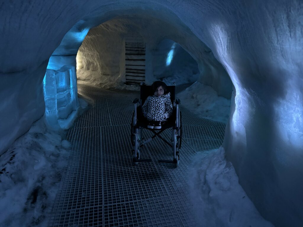 A tiny girl in a hoodie huddled in a wheelchair in an ice cave.