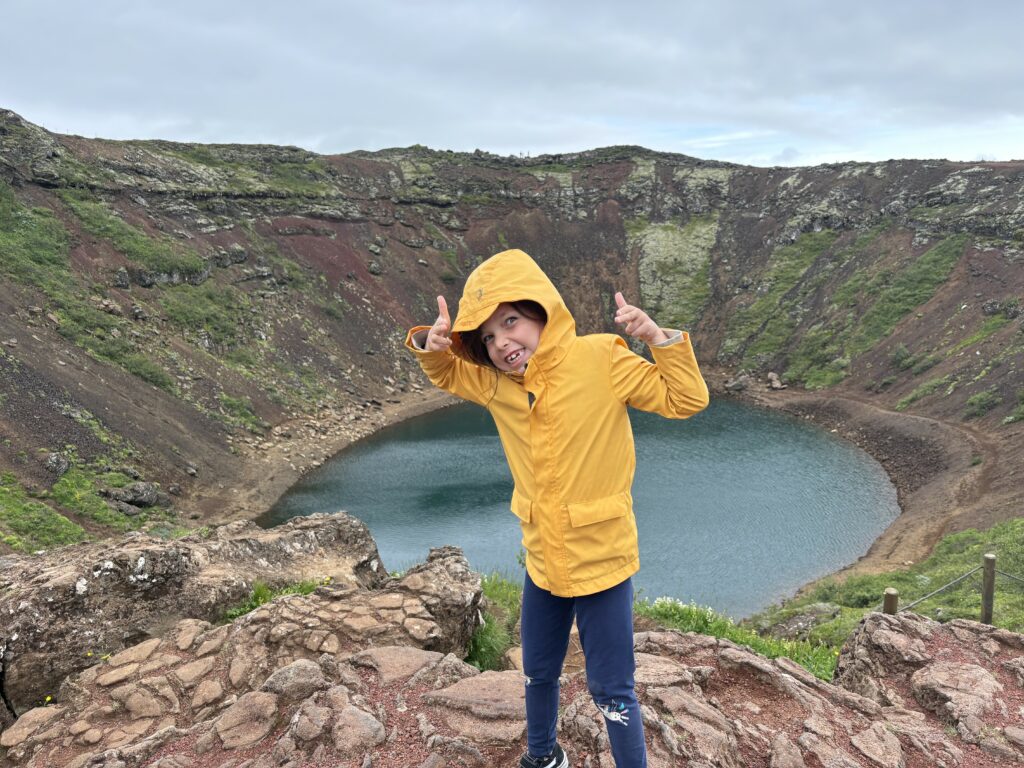 Grinning Z giving 2 thumbs up in a yellow rainjacket in front of a giant volcano crater